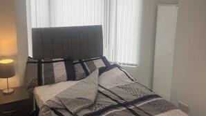 A bedroom with a bed, a bedside table with a lamp, and a window with blinds. - Stylish Dream Home With 4 En-suite Bedrooms (Liverpool)