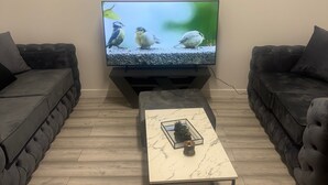 A living room with a television displaying birds, two sofas, a coffee table, and a clock on the wall. - Stylish Dream Home With 4 En-suite Bedrooms (Liverpool)