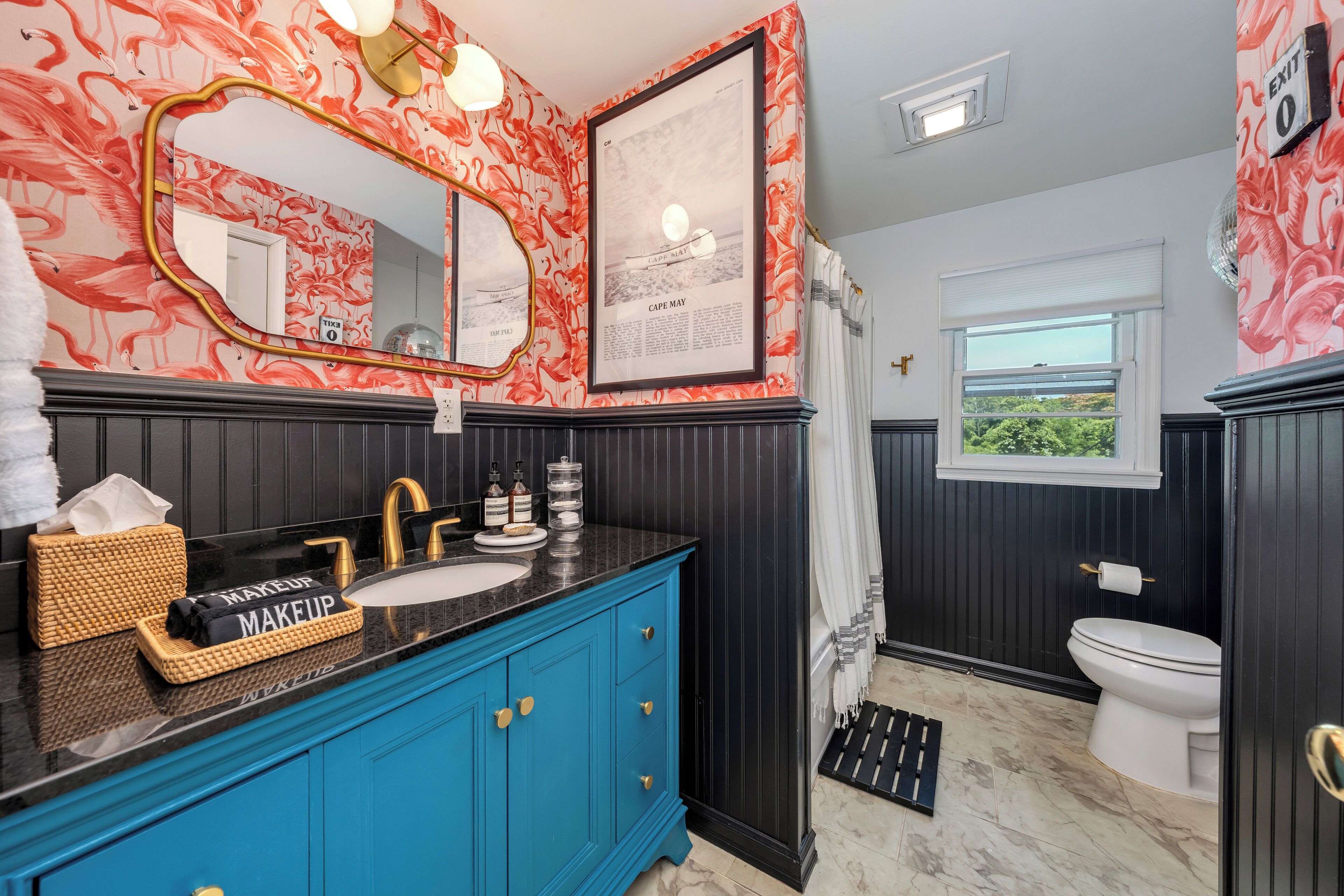 Ein Badezimmer mit blauem Waschtisch, einem Spiegel, einem gerahmten Bild, einer Toilette und einem Fenster mit Blick.