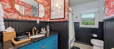 Ein Badezimmer mit blauem Waschtisch, einem Spiegel, einem gerahmten Bild, einer Toilette und einem Fenster mit Blick.