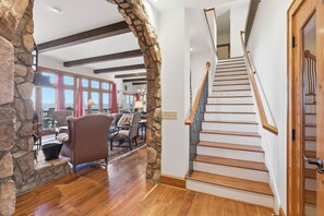 A spacious living area with a stone archway, wooden flooring, and a staircase with wooden steps and a metal railing. - LUX Lodge - Sauna, Chef Kitchen+ Long Range Views! (Banner Elk)