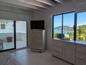 A room with a large window offering a view of the sea and hills, a television on a cabinet, and a potted plant on the windowsill. - 3-bedroom Villa with private pool and stunning views of Playa La Ropa (Zihuatanejo)