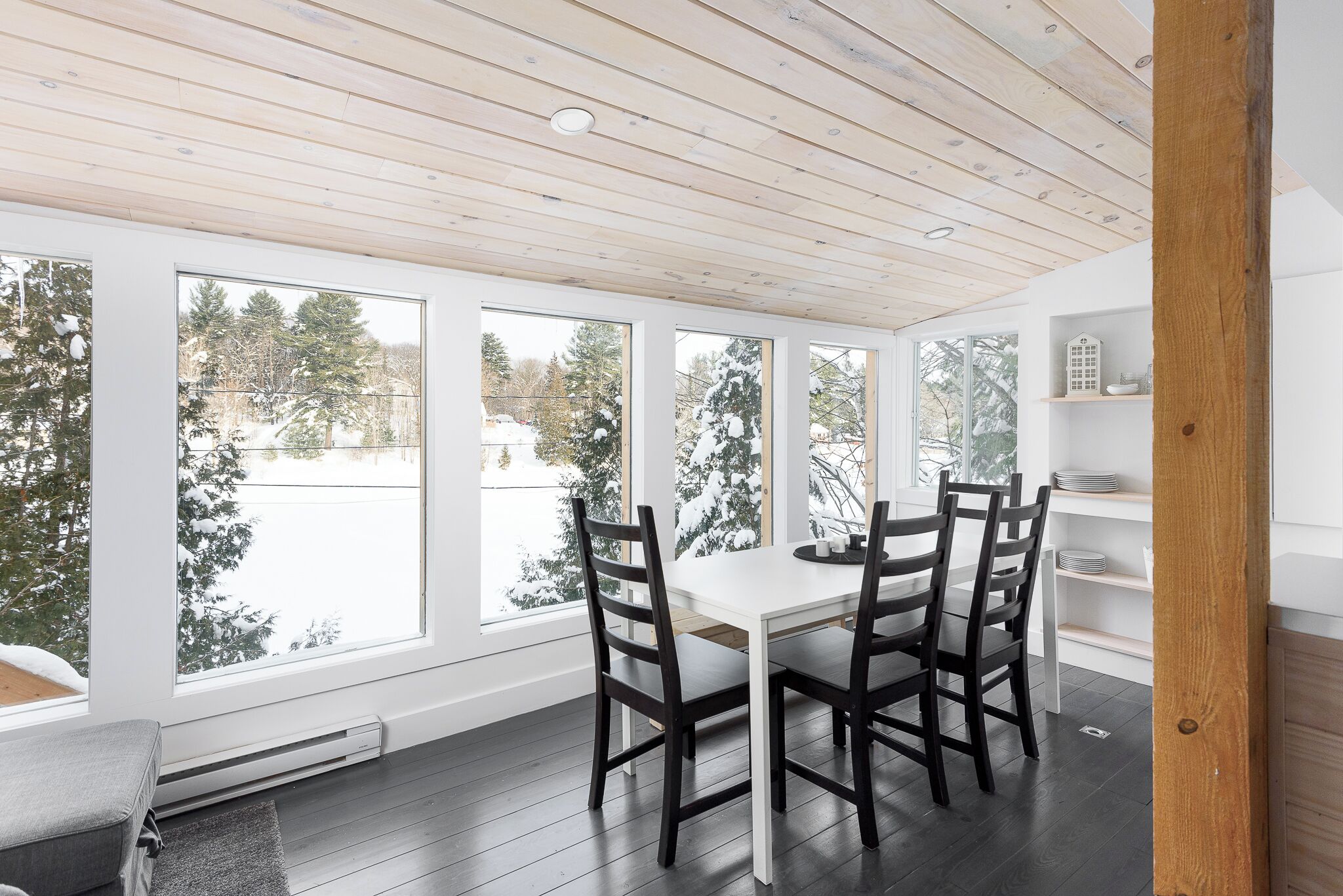 Un comedor con una mesa blanca y sillas oscuras, ventanas amplias con vistas nevadas y un techo de madera.