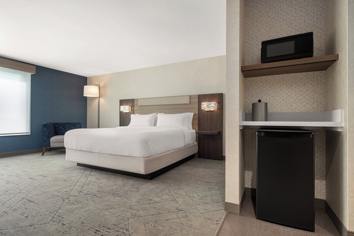 Habitación de hotel con cama, lámpara de noche, un refrigerador negro pequeño y un microondas en un estante.