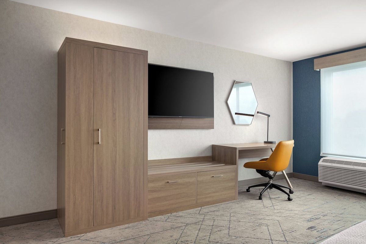 Habitación de hotel con armario, escritorio, silla, televisor de pantalla plana, espejo y ventana con unidad de aire acondicionado.