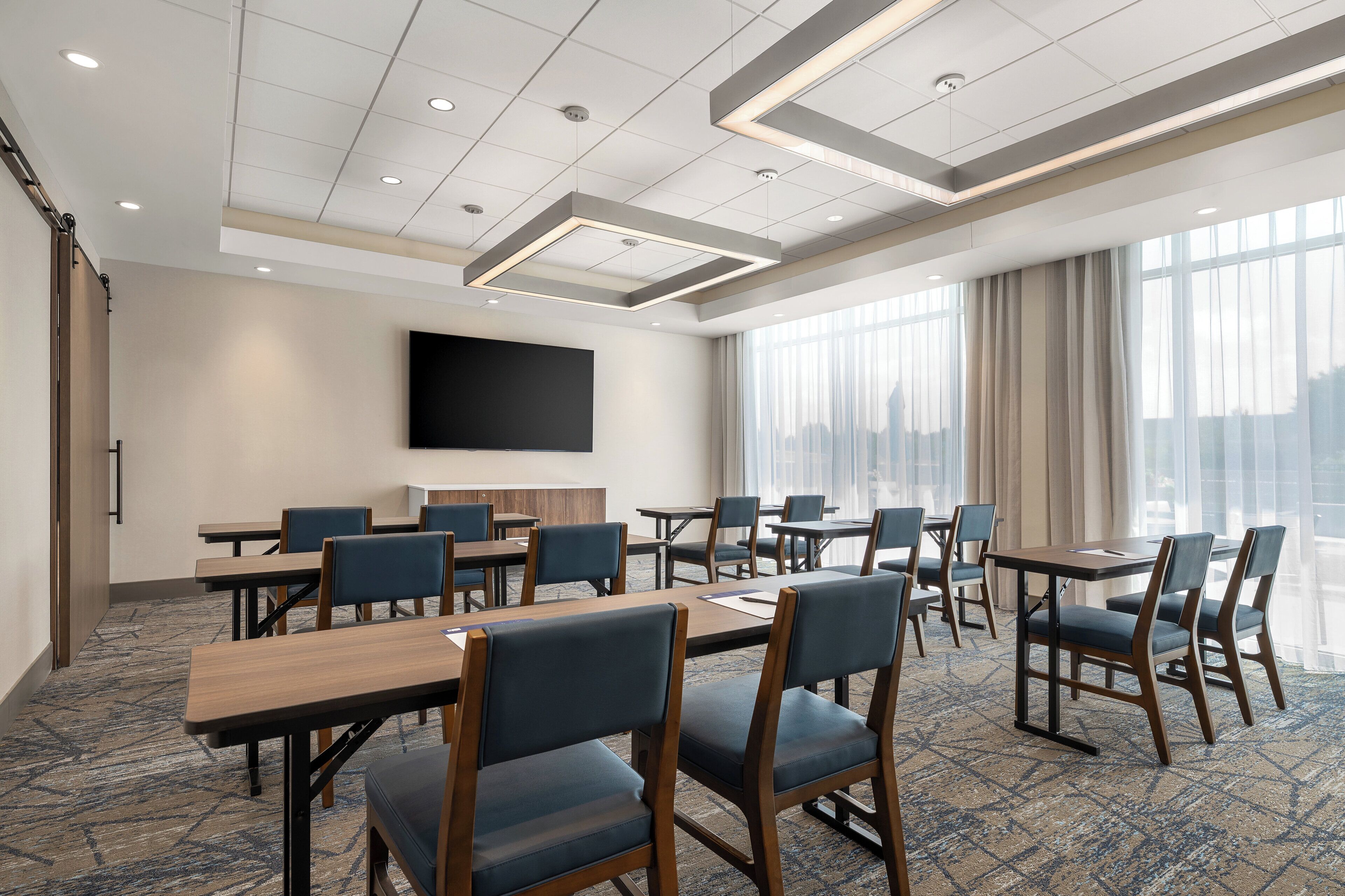 A conference room with tables and chairs, a flat-screen TV on the wall, and large windows with curtains.