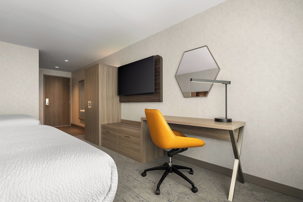 Una habitación de hotel moderna con una cama grande, un escritorio de madera, una silla de oficina naranja, un televisor de pantalla plana y un espejo hexagonal.