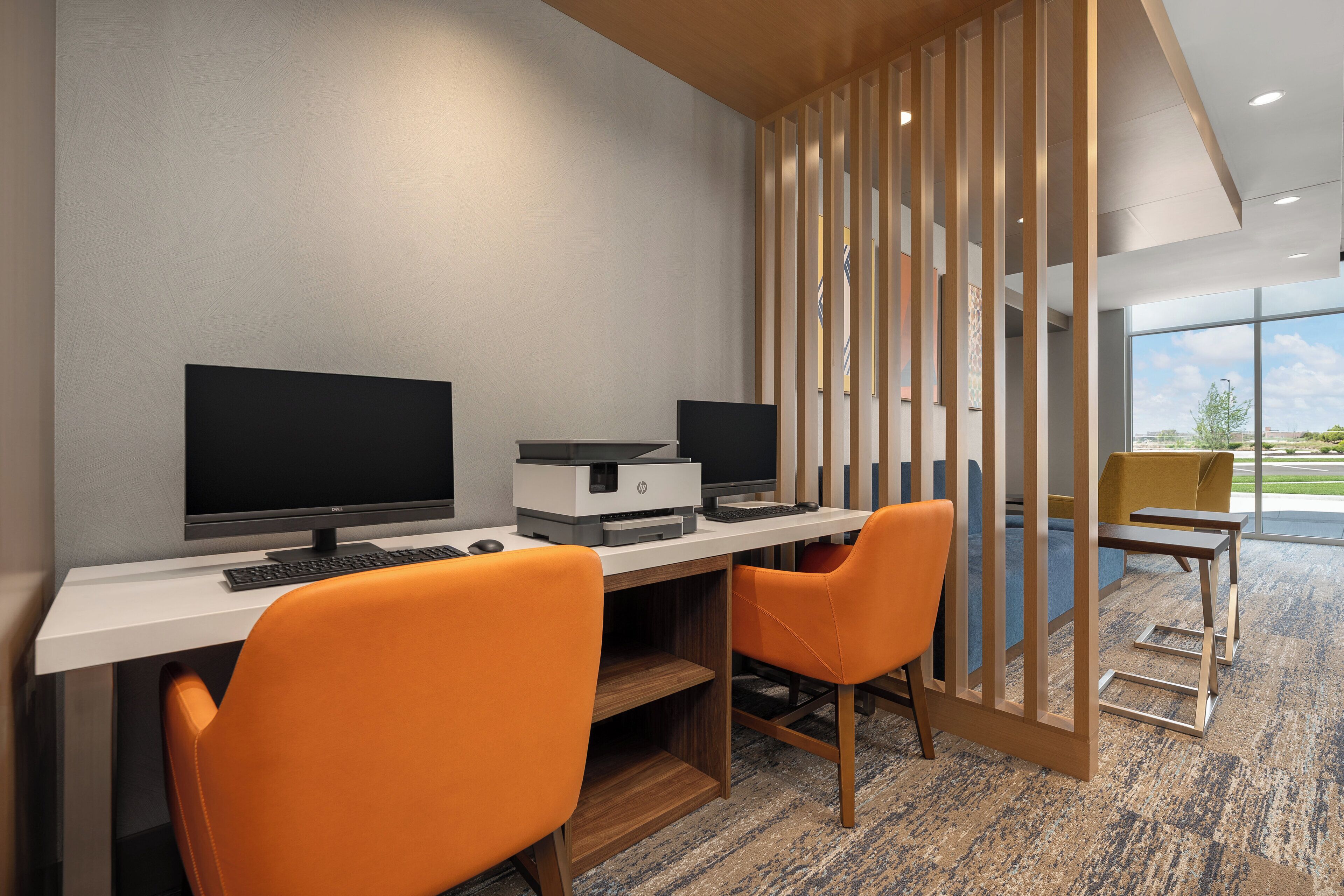 Un espace de bureau moderne avec un bureau blanc, deux chaises orange, un écran d’ordinateur, une imprimante et une grande fenêtre donnant sur l’extérieur.