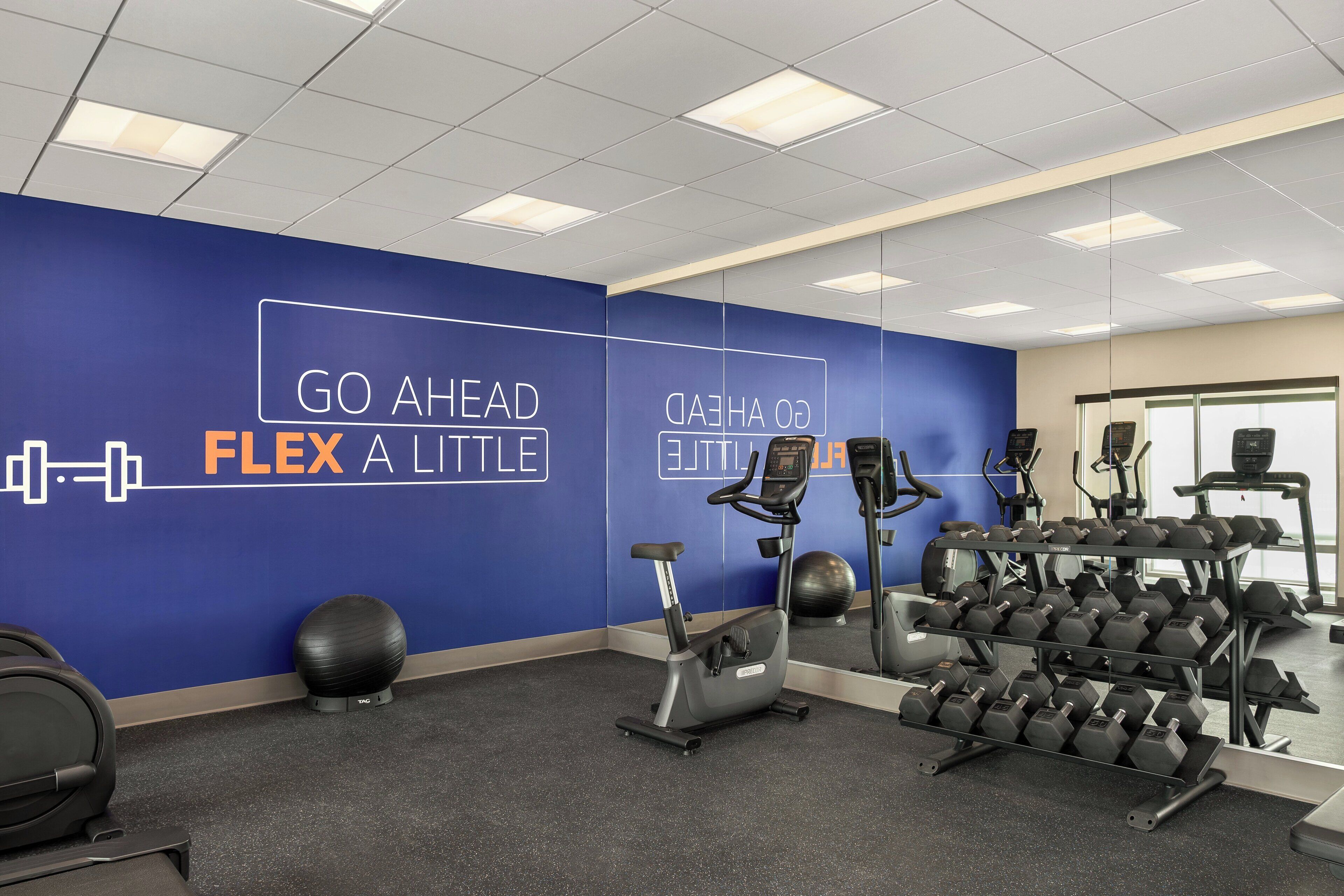 Un gimnasio con equipamiento de ejercicio, una pared con texto motivacional y un estante de mancuernas.