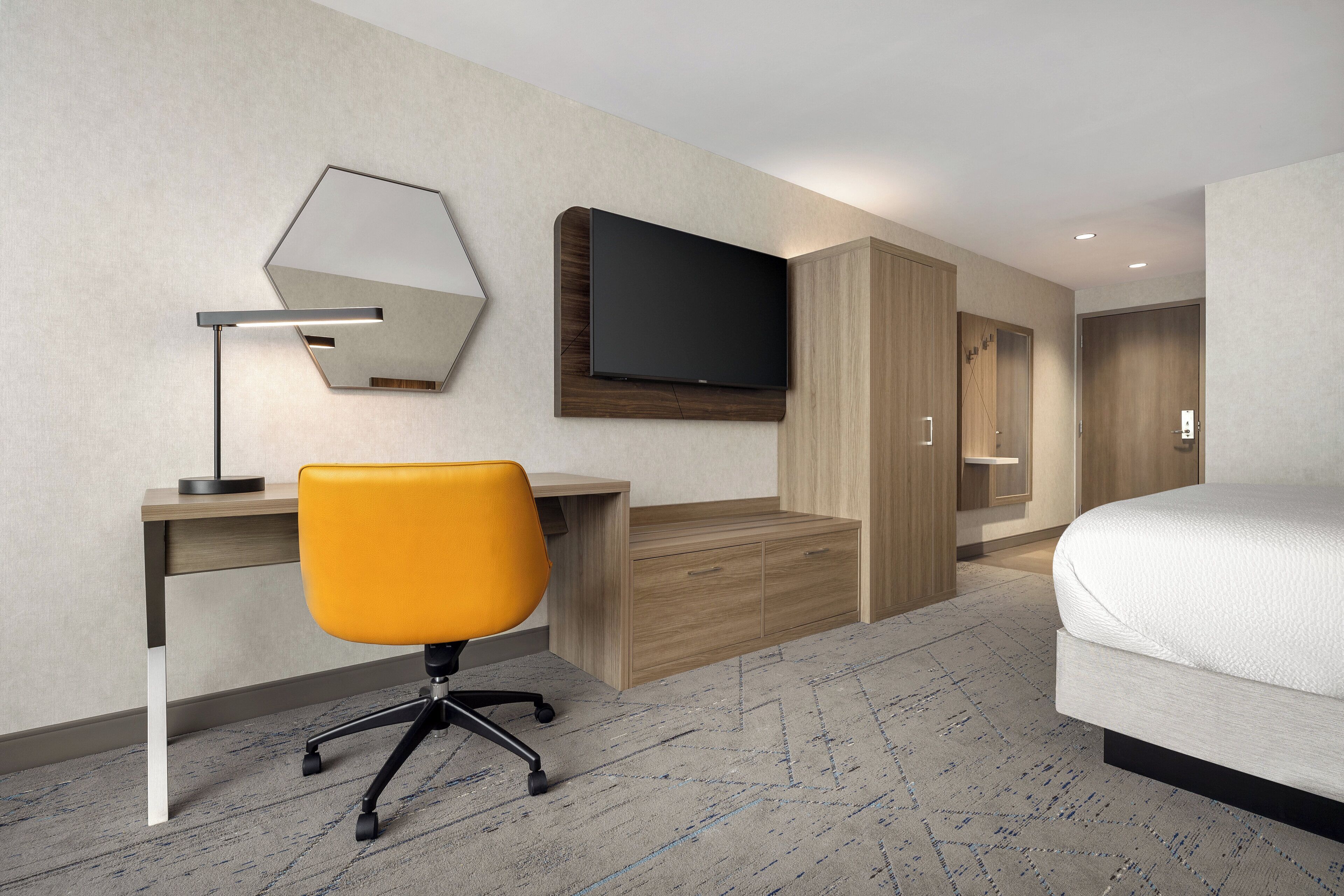 A hotel room with a bed, a desk with a chair, a TV mounted on the wall, and a wooden wardrobe.