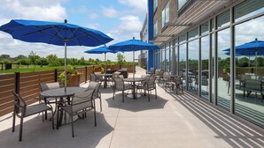 Un patio al aire libre con mesas y sillas, sombrillas azules y vista del exterior del edificio.