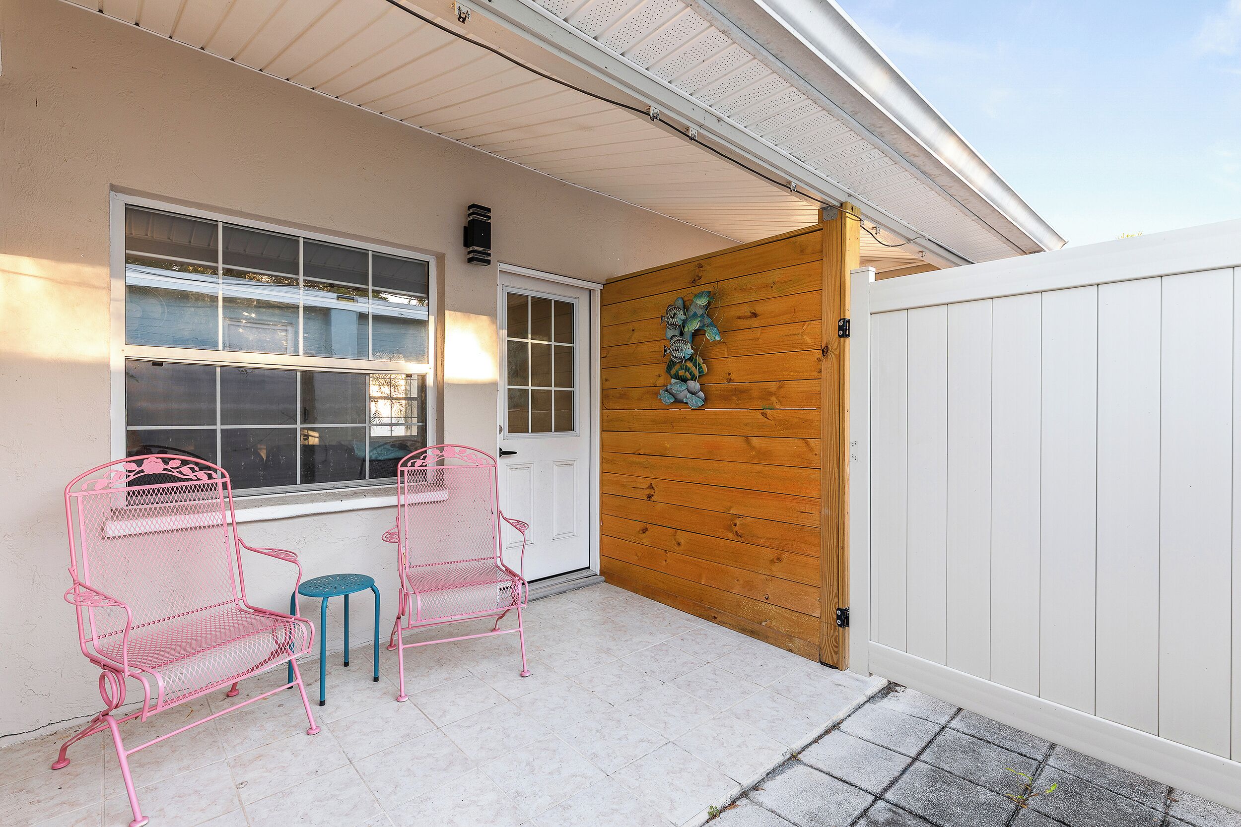 A covered patio with two pink metal chairs and a small round table, a wooden door, and a white fence.