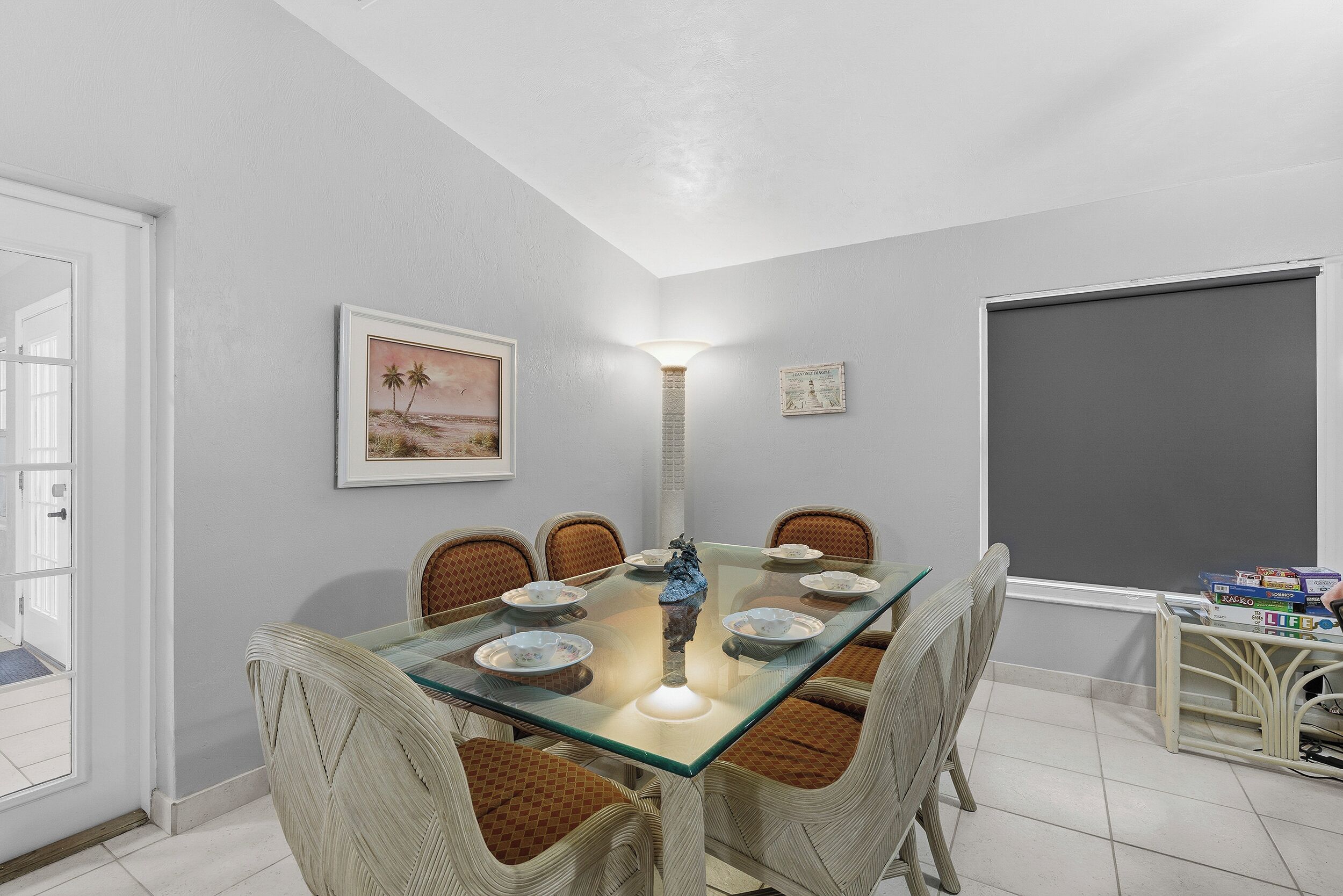 Un comedor con una mesa de vidrio, sillas de mimbre, una lámpara de pared y un cuadro enmarcado.