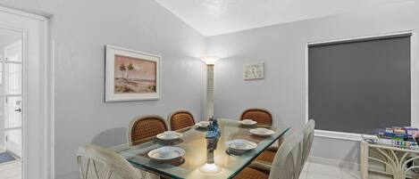 Un comedor con una mesa de vidrio, sillas de mimbre, una lámpara de pared y un cuadro enmarcado.