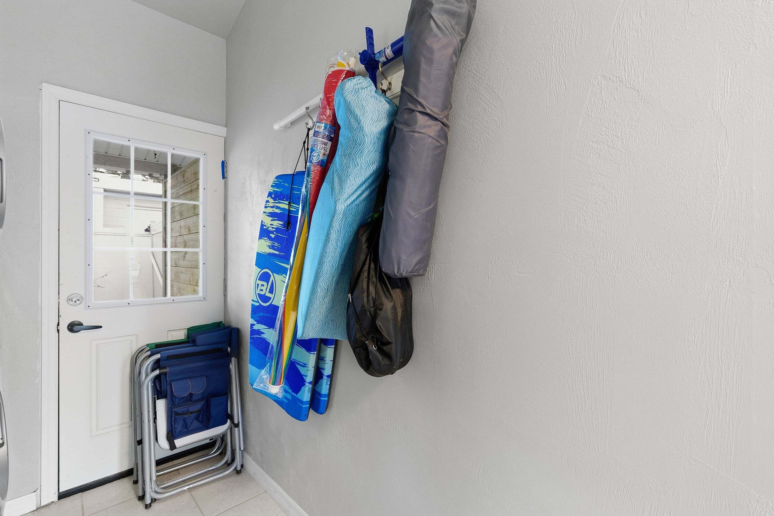 Ein Zimmer mit einer weißen Tür, einem Fenster, einer blauen Matte, einer gefalteten blauen Decke, einer schwarzen Tasche und einem grauen Kissen an der Wand.
