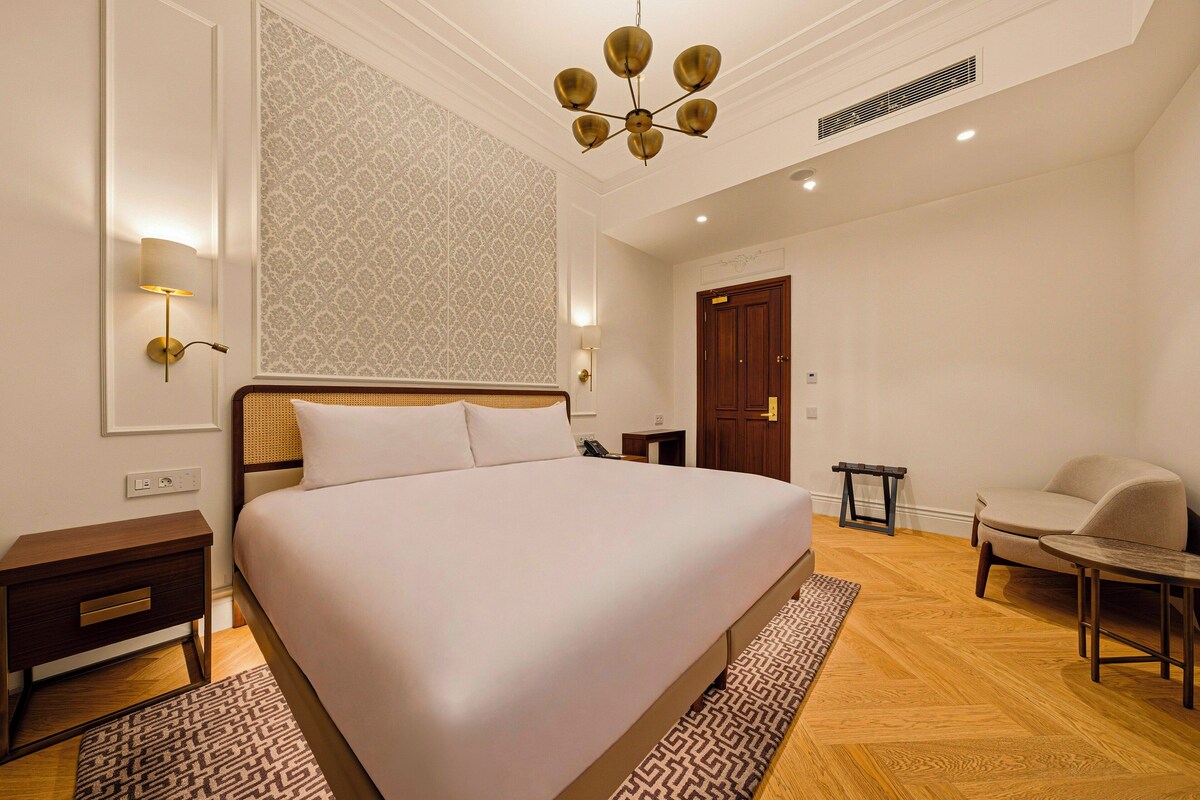 A hotel room with a large bed, bedside tables, a chair, a small table, a patterned rug, and a ceiling light fixture.