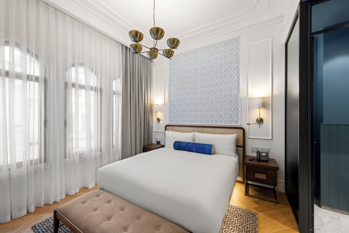 Hotellihuone, jossa on suuri sänky, yöpöytä lampulla, seinään kiinnitetty puhelin ja ikkuna, jossa on ohuet verhot.