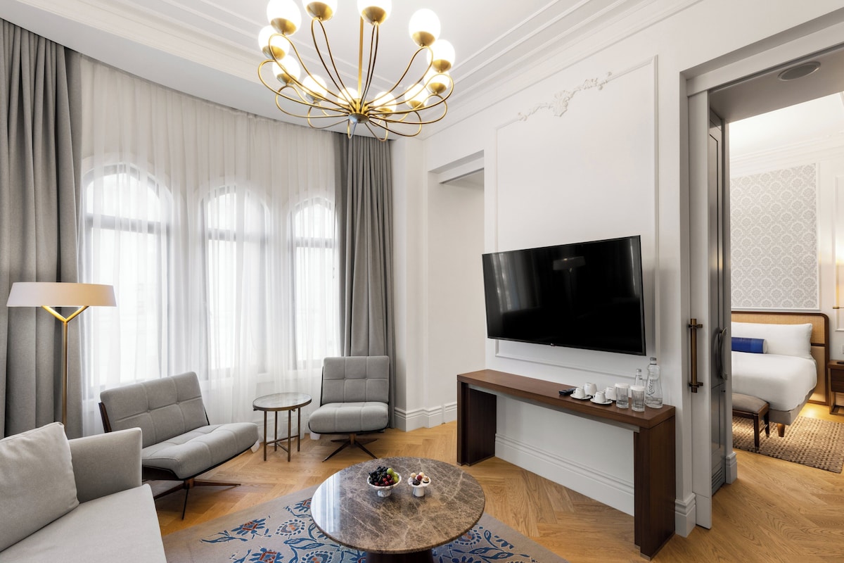 Moderni hotellihuone, jossa on litteänäyttöinen televisio, harmaalla kankaalla verhoiltu istuinryhmä, pyöreä sohvapöytä, jossa on hedelmiä, ja koristeellinen kattokruunu.