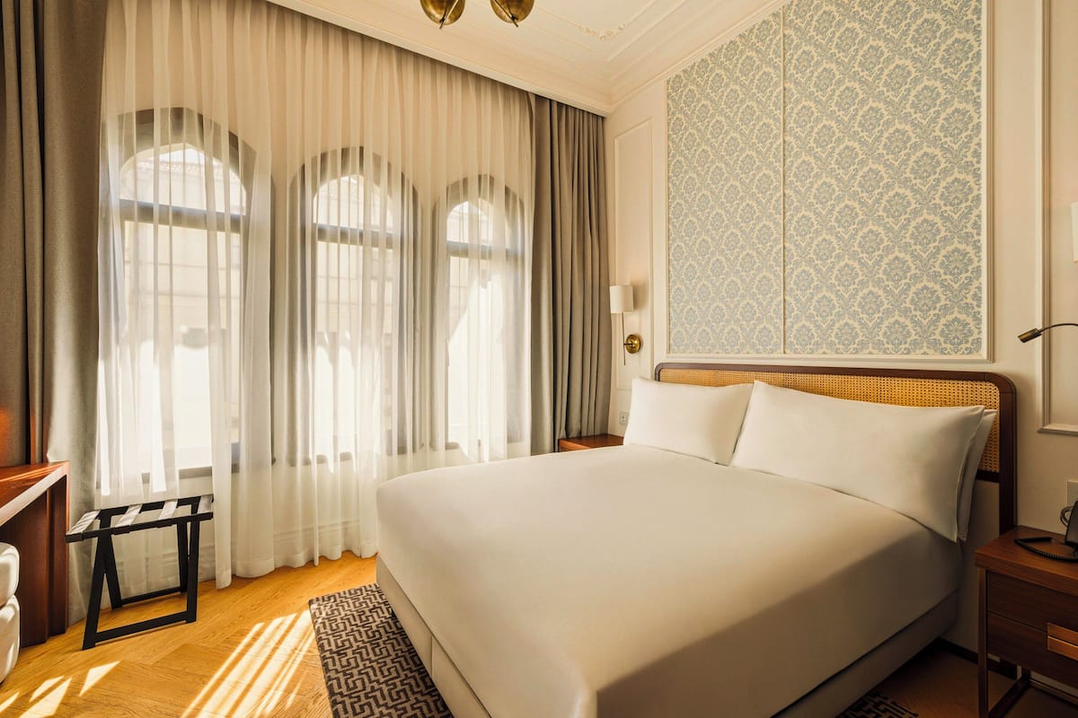 A hotel room with a large bed, a bedside table, a nightstand, a lamp, and a window with curtains.