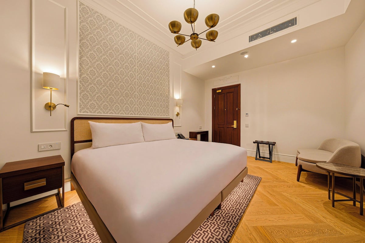A hotel room with a large bed, bedside tables, a chair, a small table, a patterned rug, and a ceiling light fixture.