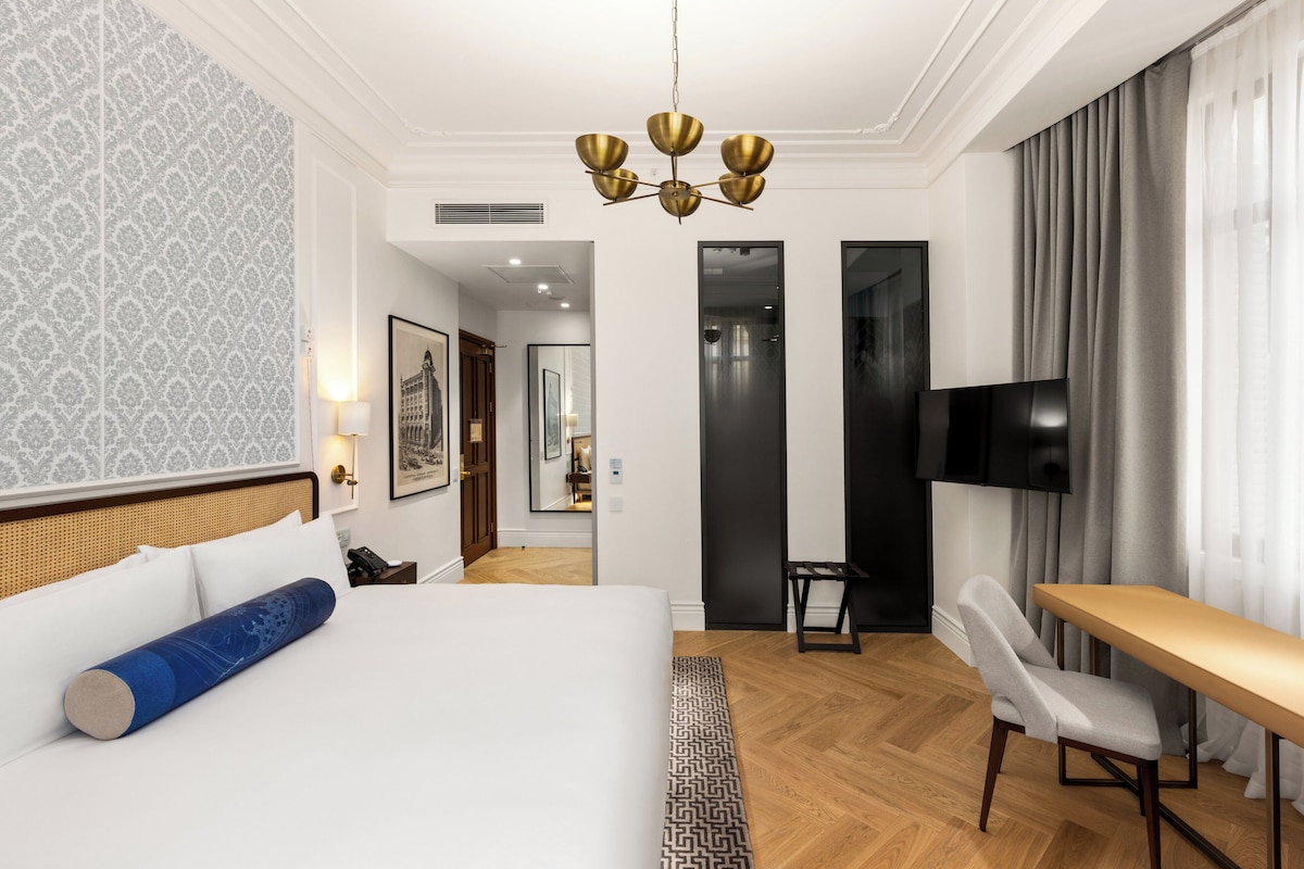 Hotellihuone, jossa on sänky, työpöytä, tuoli, televisio ja käytävä, joka johtaa toiseen huoneeseen.