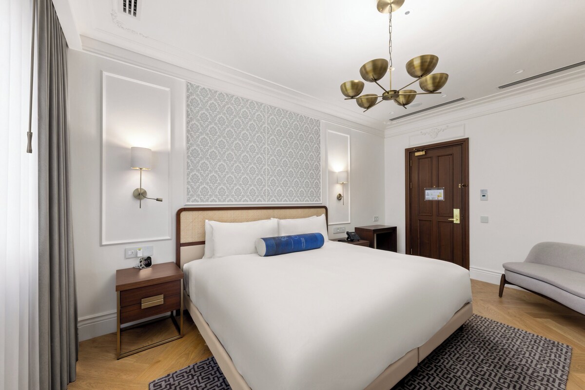 Hotellihuone, jossa on suuri sänky, yöpöydät, työpöytä, tuoli, kuvioitu matto, kattokruunu ja ovi.