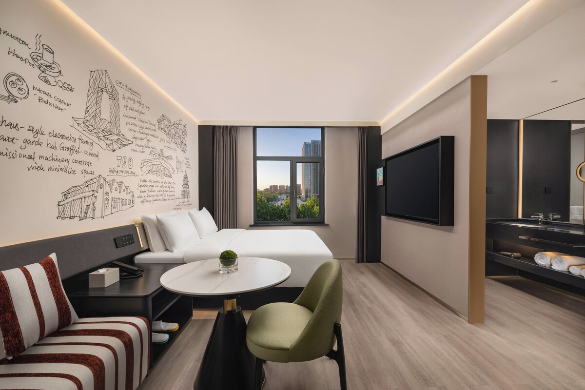 A modern hotel room with a bed, a sofa, a dining table, and a TV.
