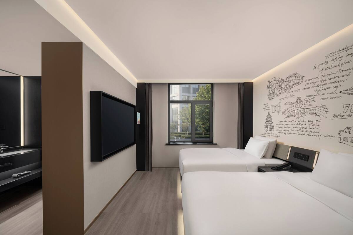Ein modernes Hotelzimmer mit einem großen Flachbildfernseher, zwei Betten, einem Fenster mit Blick auf die Stadt und einer Wand mit architektonischen Skizzen.