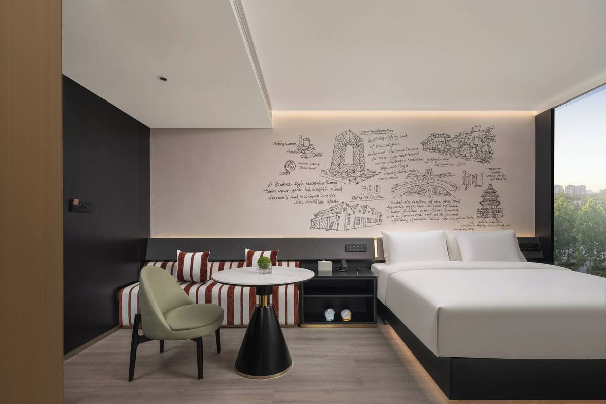 Ein modernes Hotelzimmer mit einem großen Bett, einer Sitzecke mit gestreiften Kissen, einem kleinen Tisch mit einer Pflanze und einer Wand mit architektonischen Skizzen.