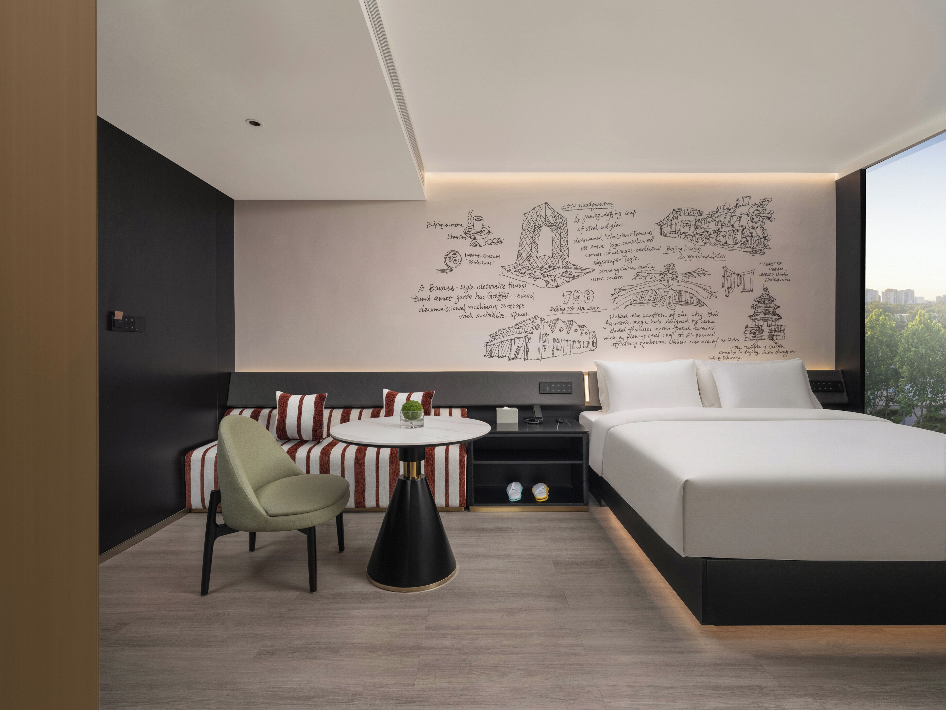 Une chambre d’hôtel moderne avec un grand lit, un coin salon avec des coussins à rayures, une petite table avec une plante et un mur orné de croquis architecturaux.