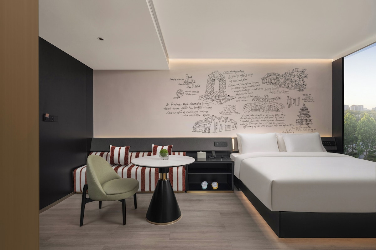 A modern hotel room with a large bed, a seating area with striped cushions, a small table with a plant, and a wall with architectural sketches.