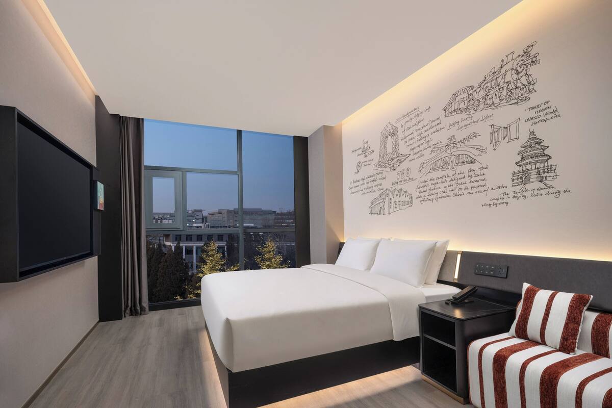 Ein modernes Hotelzimmer mit einem großen Bett, einem Nachttisch, einem Fernseher und Blick auf eine Stadtlandschaft durch große Fenster.
