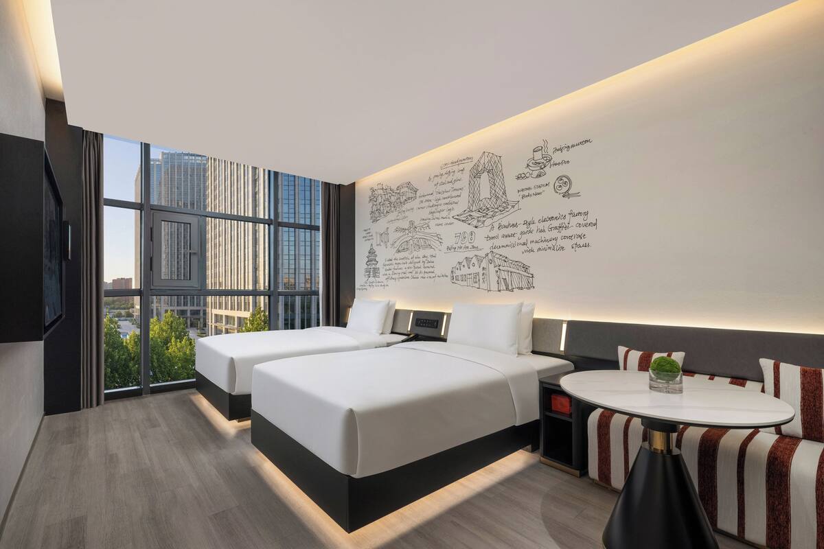 Ein modernes Hotelzimmer mit großem Fenster, zwei Betten, einem kleinen Tisch und einer Wand mit Skizzen.