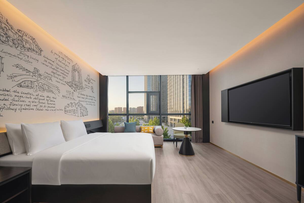 Ein modernes Hotelzimmer mit einem großen Bett, einem Schreibtisch, einem Sessel, einem kleinen Tisch und einem großen Fenster mit Blick auf die Stadt.