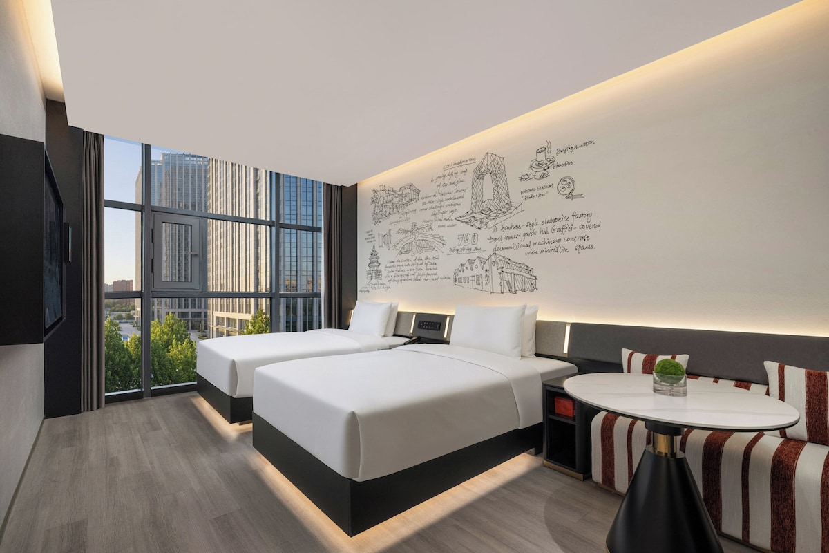 A modern hotel room with a large window, two beds, a small table, and a wall with sketches.