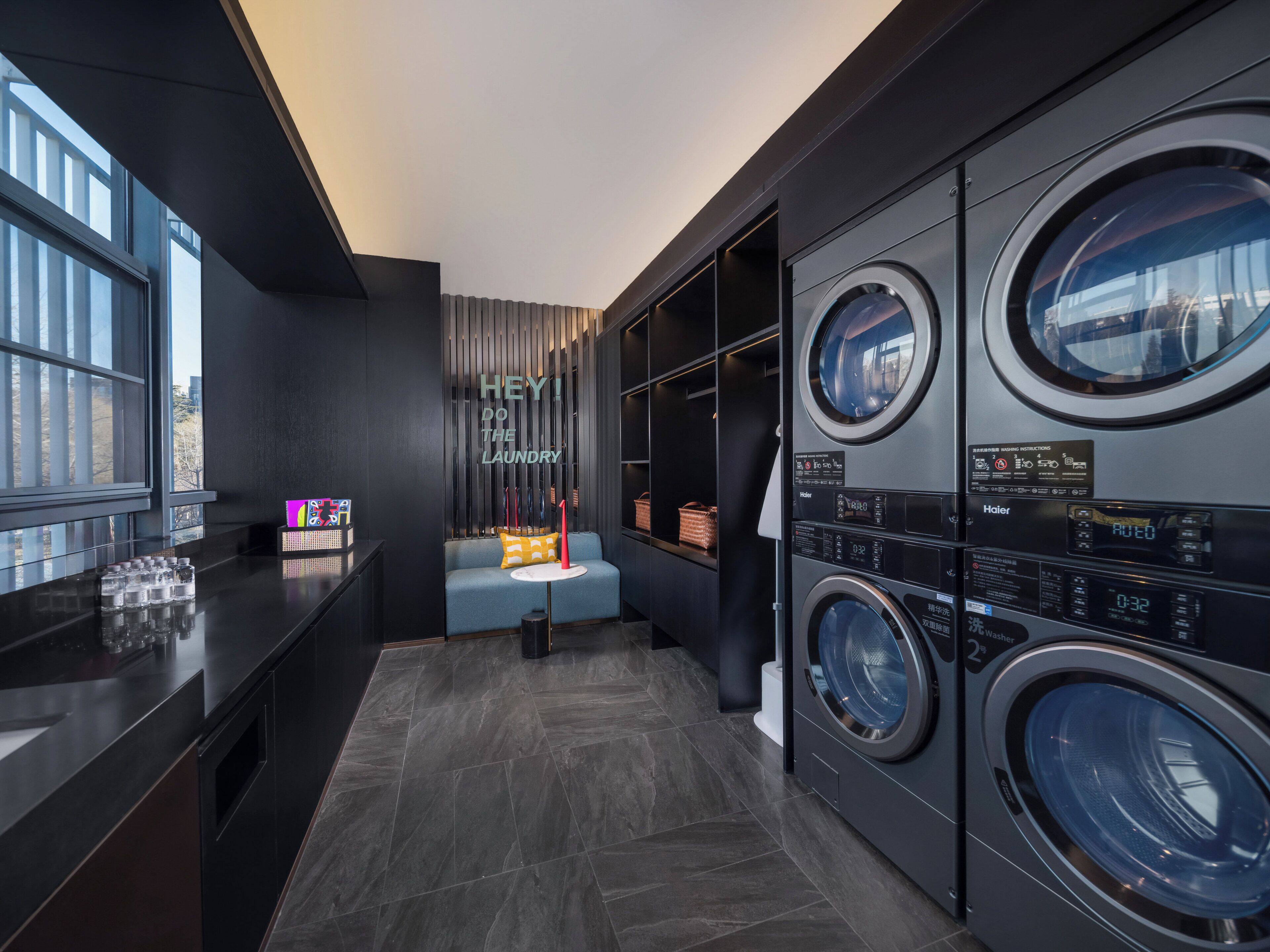 A modern laundry room with built-in washers and dryers, a seating area, and a view of the city through large windows.