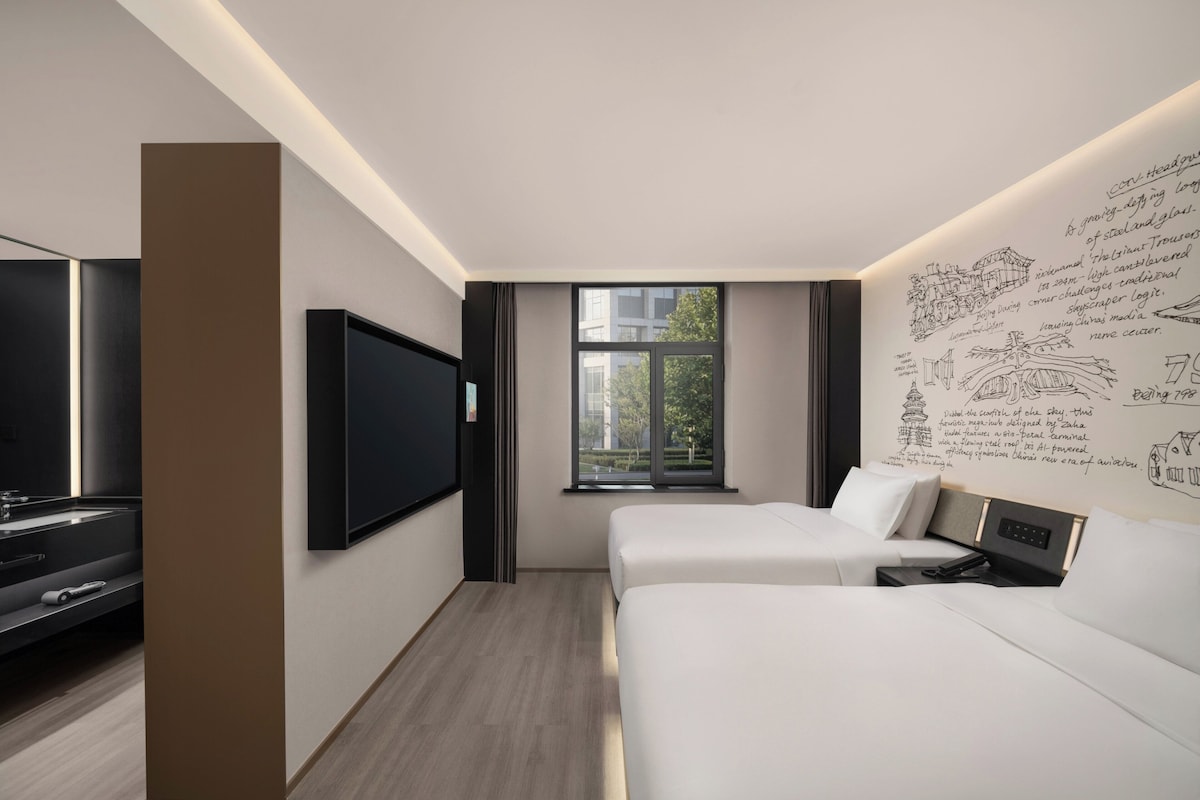 A modern hotel room with a large flat-screen TV, two beds, a window with a view of the city, and a wall with architectural sketches.