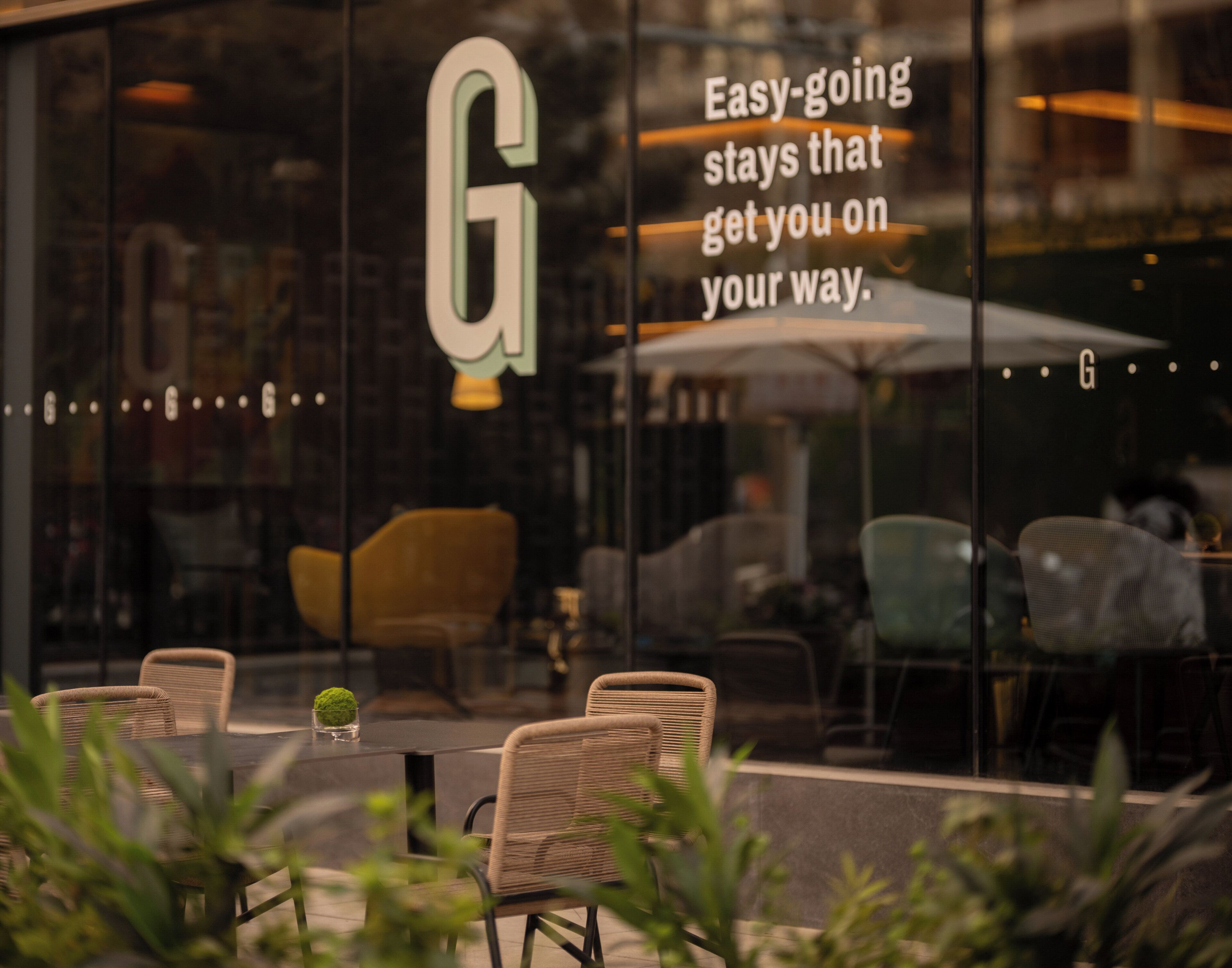 A restaurant with outdoor seating, a large glass window with a 'G' logo, and a quote about easy-going stays.