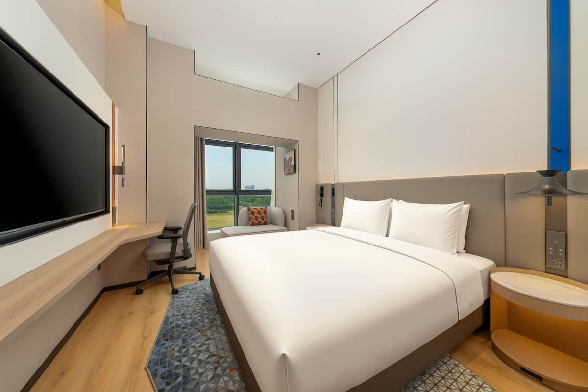 A modern hotel room with a large bed, a desk, a chair, a TV, and a window with a view.