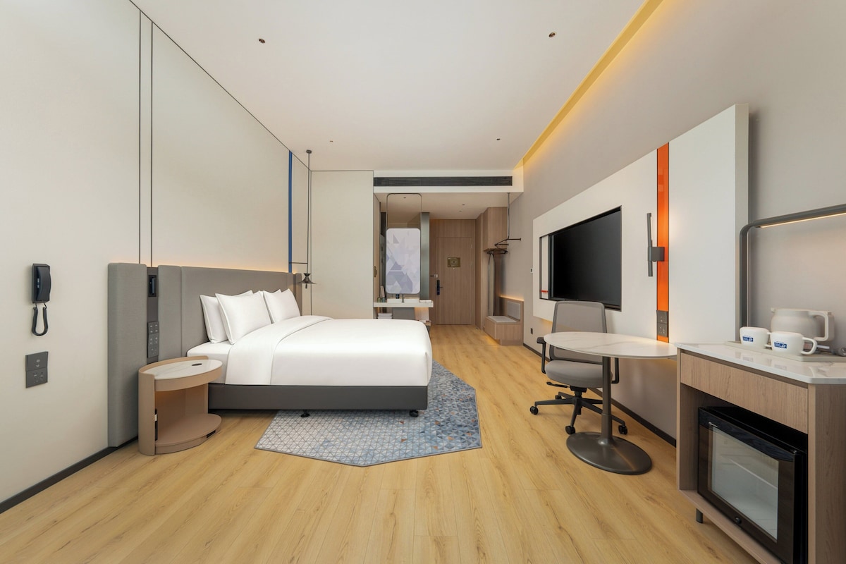 A modern hotel room with a bed, bedside table, desk, chair, TV, and a small kitchenette.