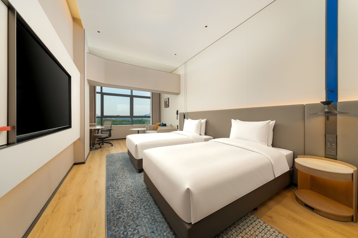 A modern hotel room with two beds, a large window, a flat-screen TV, and a small desk.