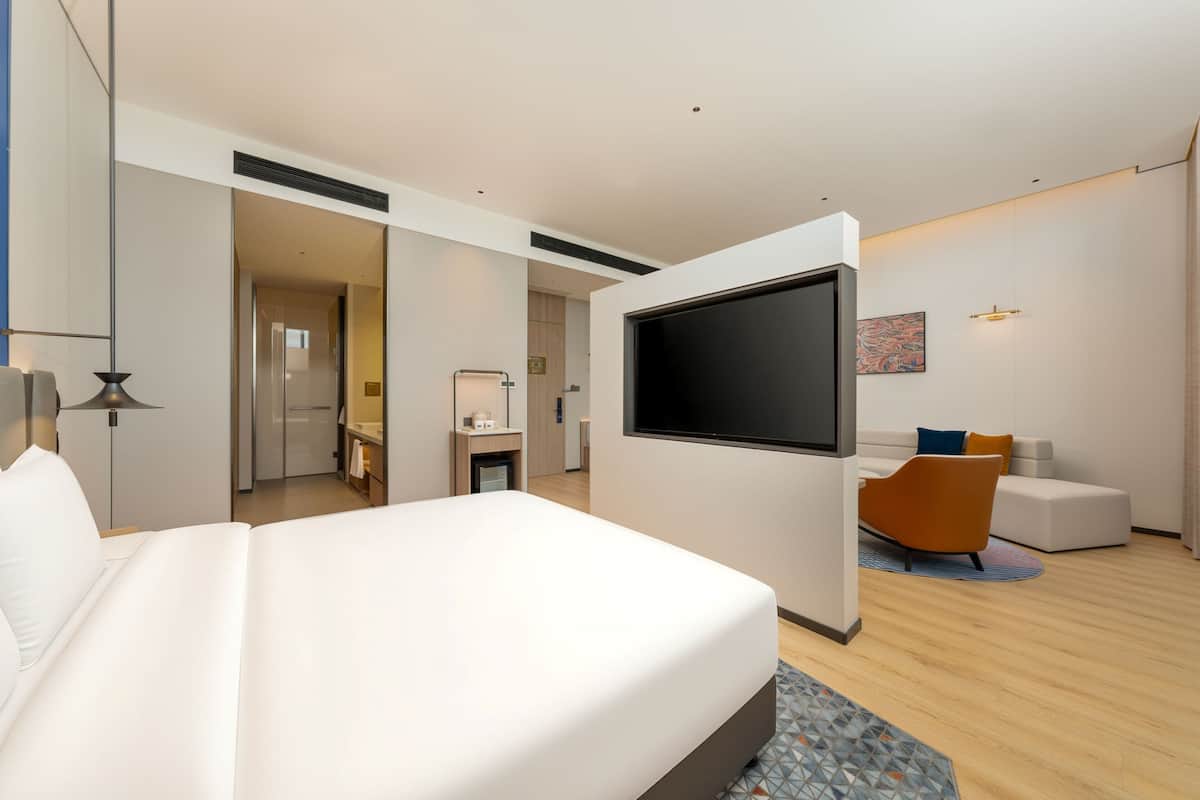 A modern hotel room with a large bed, a TV, a sofa, and a bathroom visible through an open door.