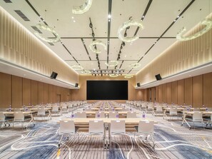 Un salón de conferencias con una pantalla grande, varias mesas con sillas e iluminación moderna en el techo.