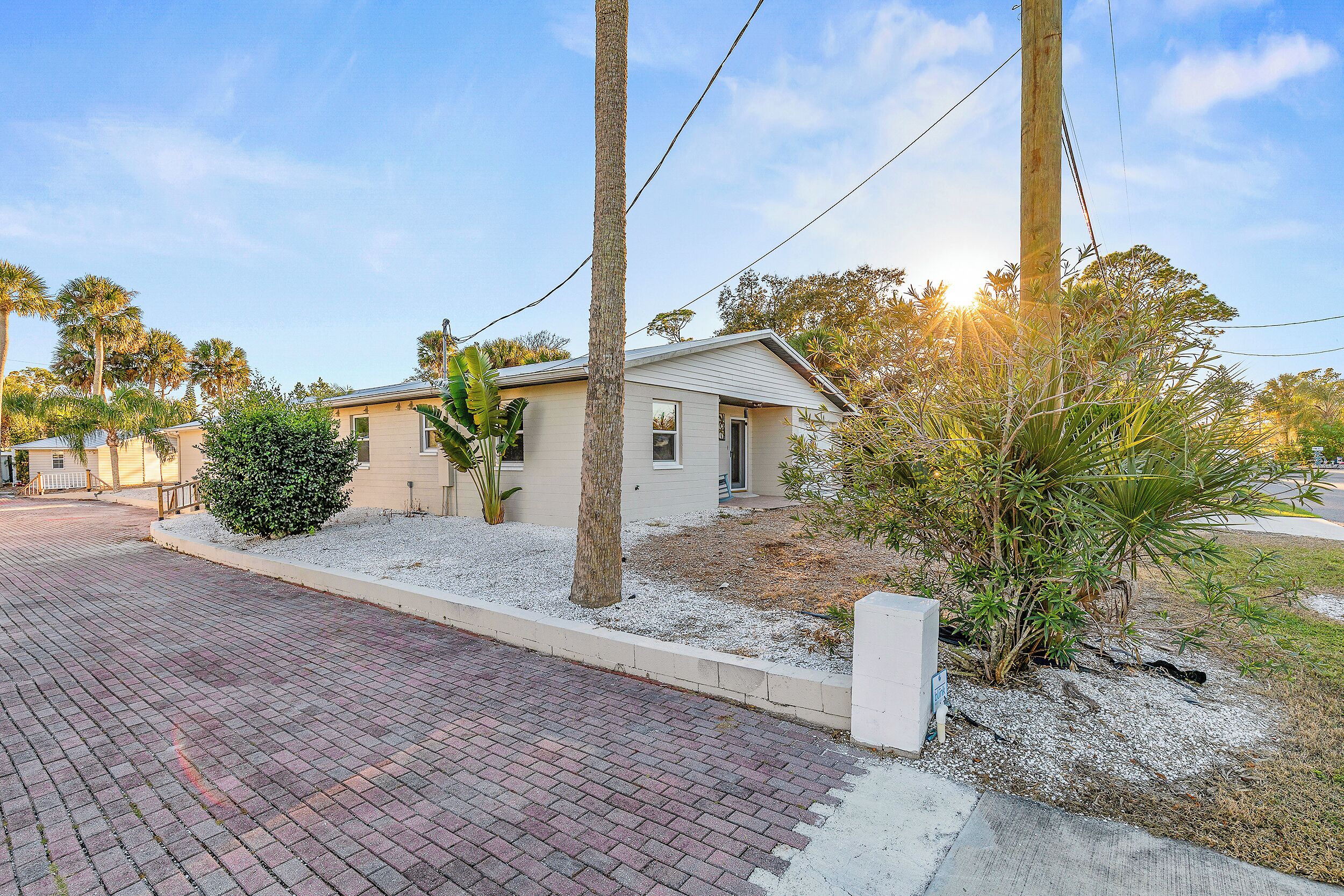 A residential property with a paved driveway, a small house with a light-colored exterior, and surrounding vegetation including palm trees and shrubs.
