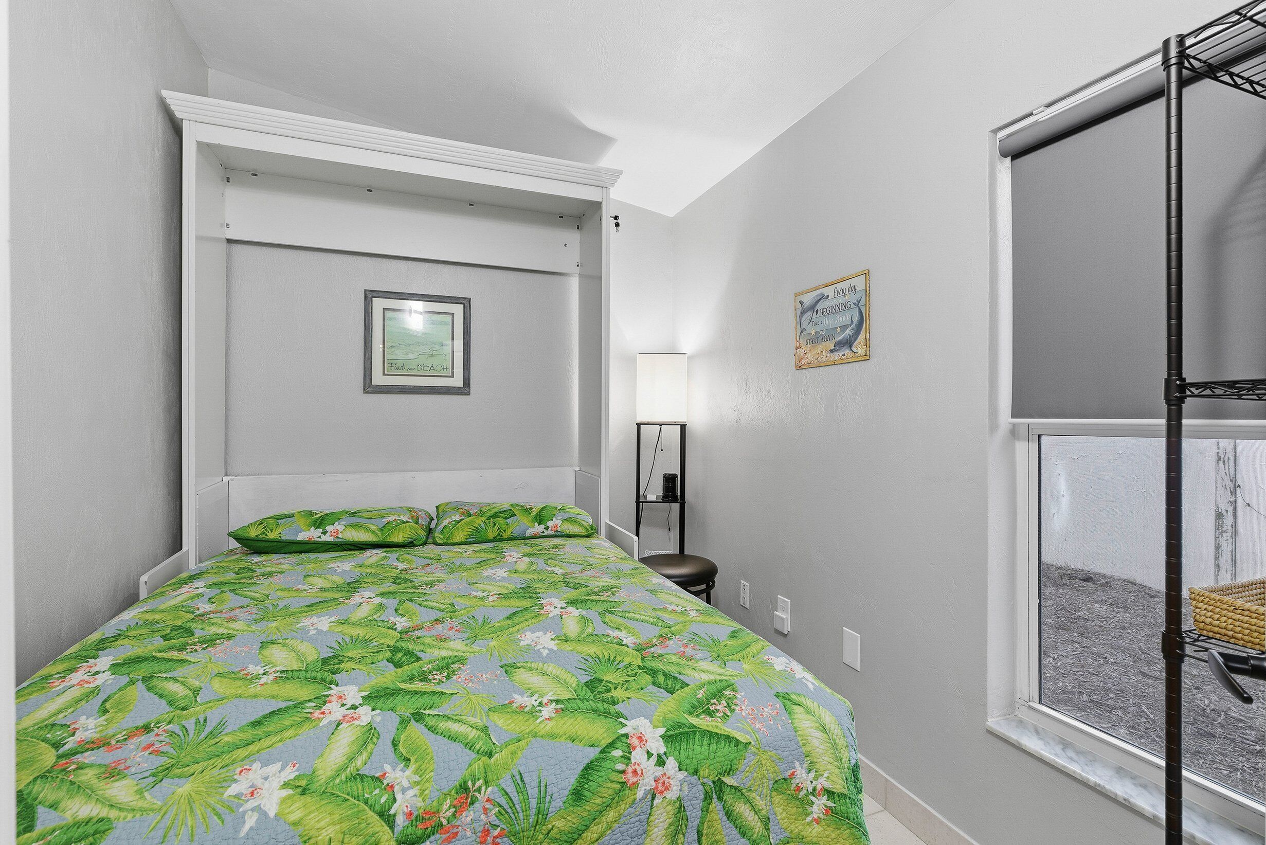 A bedroom with a bed, a window, a framed picture on the wall, and a shelf with a lamp.