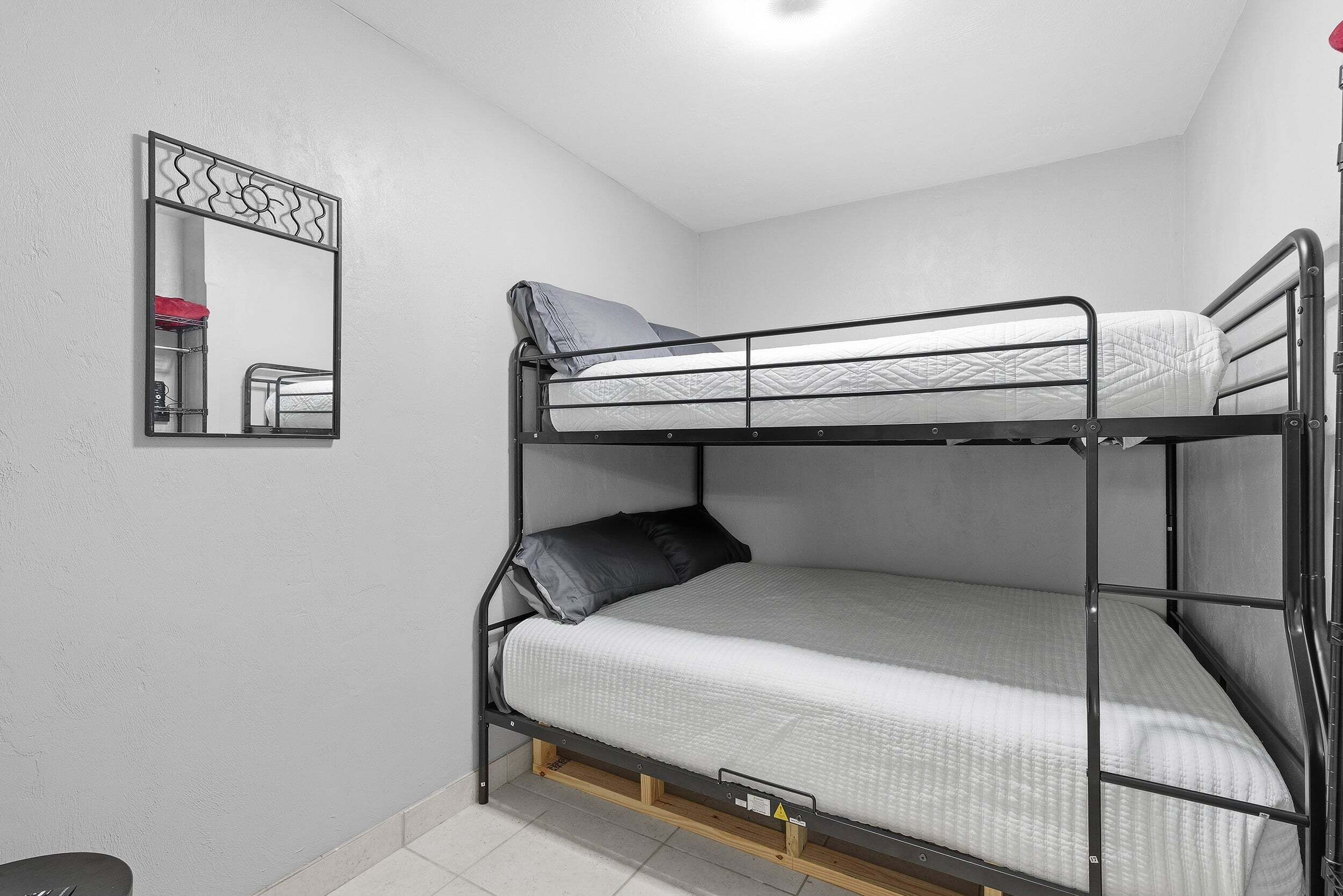Ein Etagenbett mit Spiegel und einem kleinen Regal in einem Zimmer mit weißen Wänden und Fliesenboden.