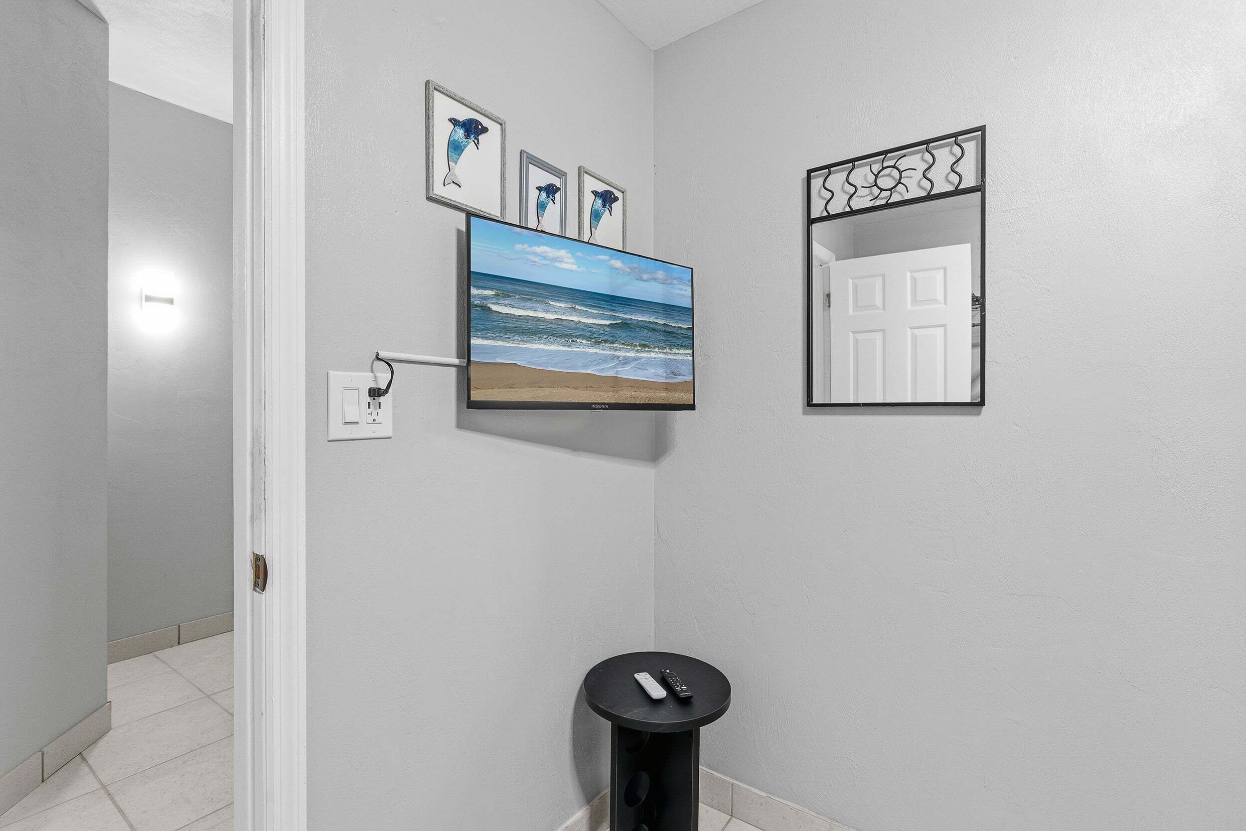 A hallway with a mounted TV displaying a beach scene, a door with a decorative mirror, and a small black table with a remote control.