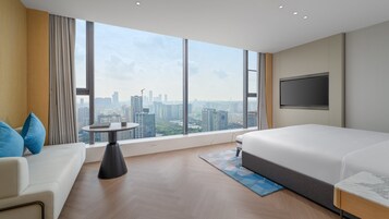 Una habitación de hotel con un ventanal grande, una cama, un televisor, una mesita pequeña y un sofá.