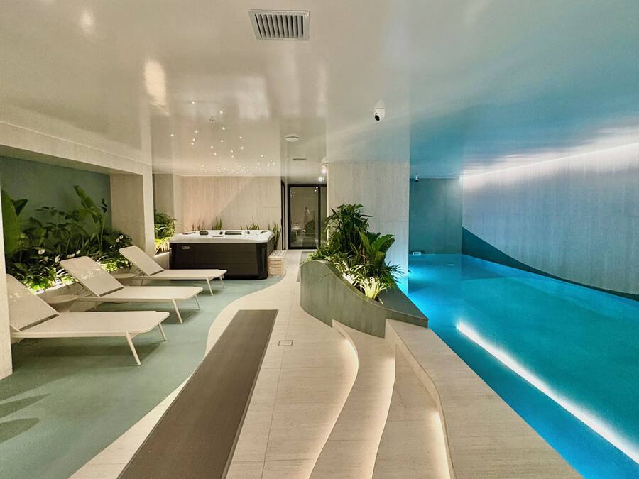 A modern indoor pool area with lounge chairs, a central seating area, and a blue-lit water feature.