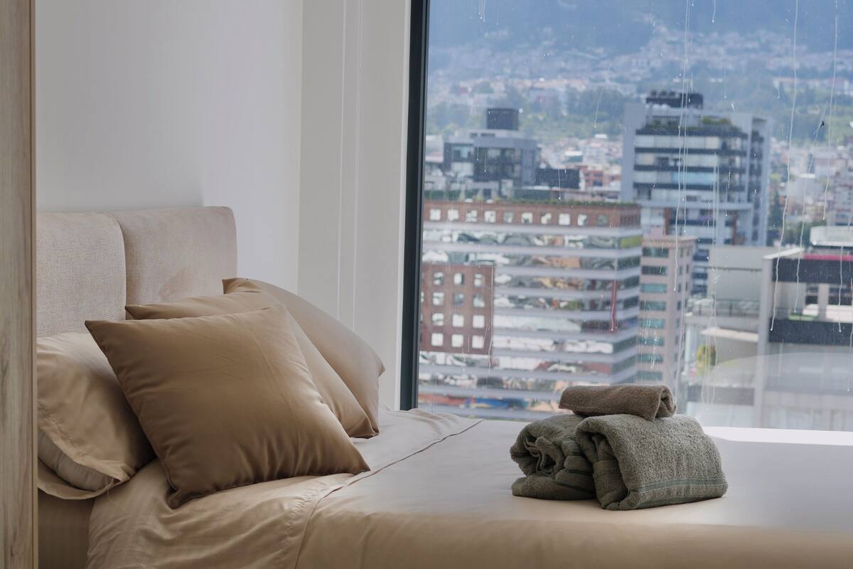 Un dormitorio con cama, almohadas, ventana con vistas a la ciudad y una toalla sobre la cama.